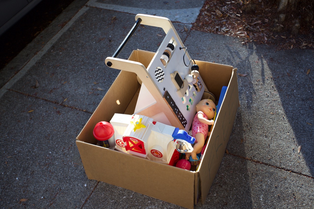 A cardboard box filled with assorted children's toys is placed on a sunlit concrete sidewalk.