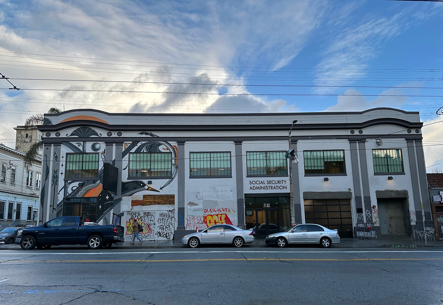 A street view of a building with a "Social Security Administration" sign, featuring murals and graffiti, with parked cars in front.