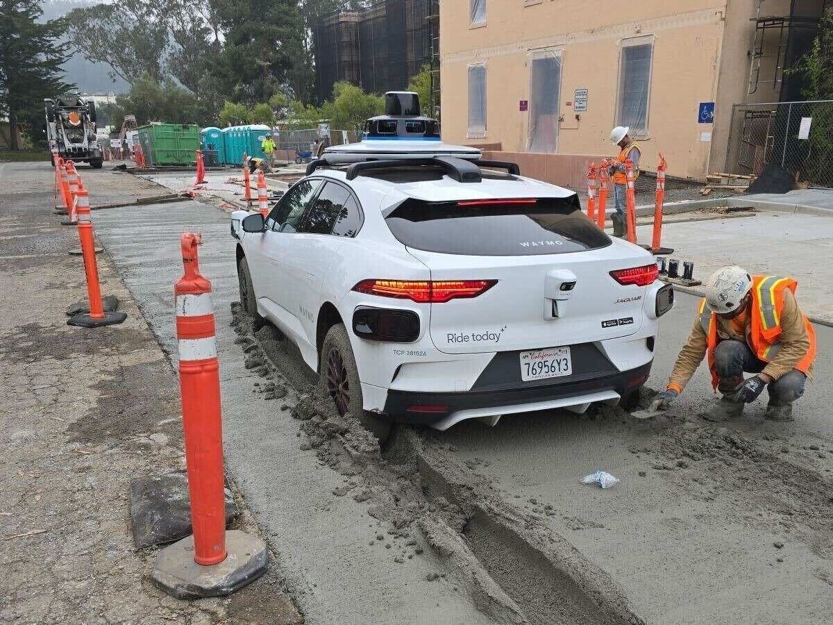 A white Waymo car is stuck in wet concrete on a construction site, surrounded by orange cones. A worker in a safety vest and helmet attempts to clear the concrete around the wheels.
