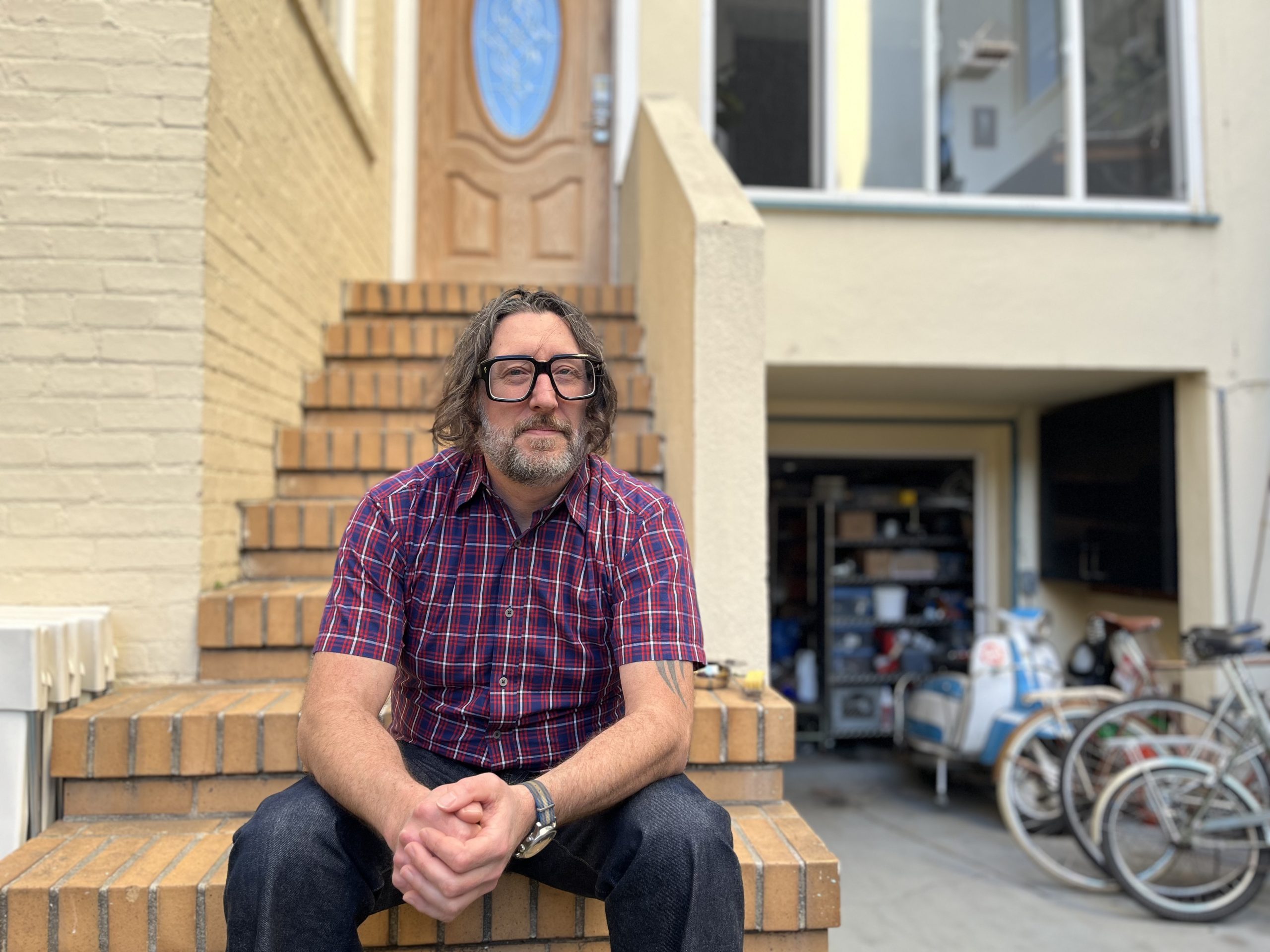A man with glasses and long hair sits on brick steps, wearing a red plaid shirt. A house with an open garage full of items is in the background.