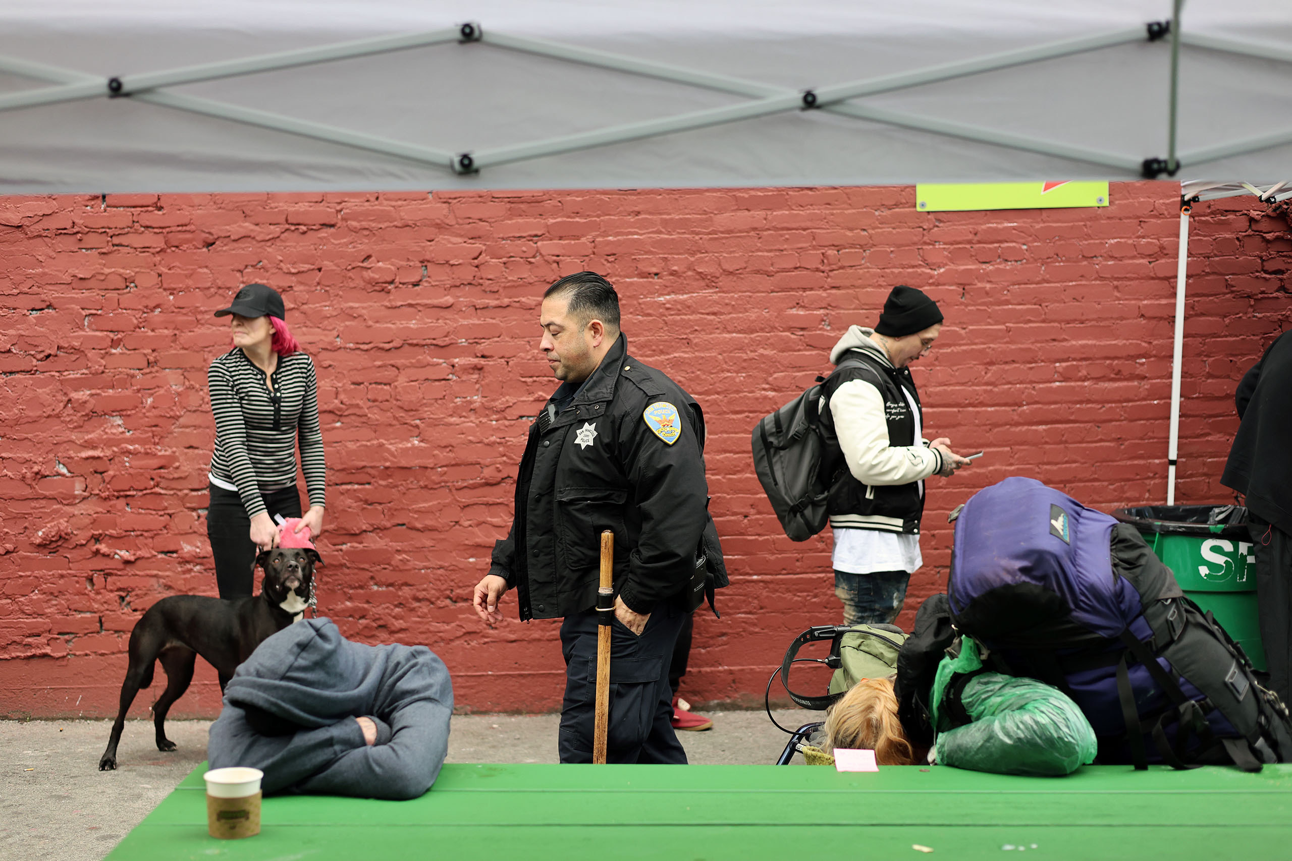 People at an outdoor area: a woman with a dog, an officer walking, a person texting, and another resting on a green table under a canopy. A brick wall serves as the background.
