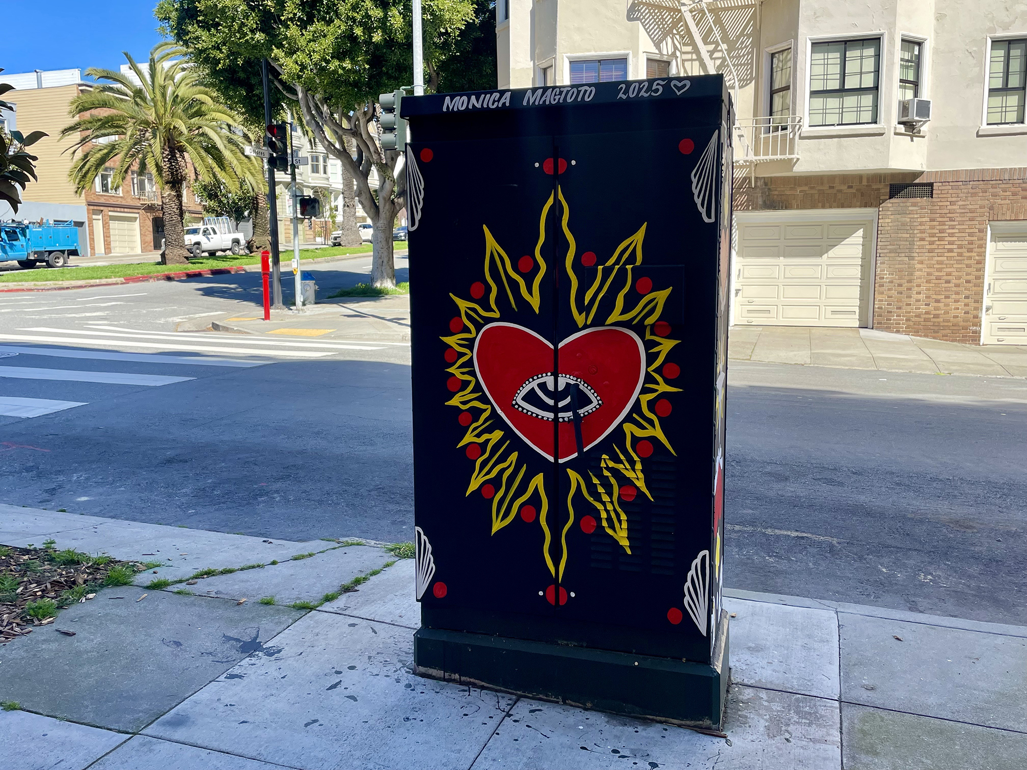 A utility box painted with a red heart, an eye in the center, surrounded by yellow rays and red teardrop shapes; "Monica Magoto 2025" is written at the top.