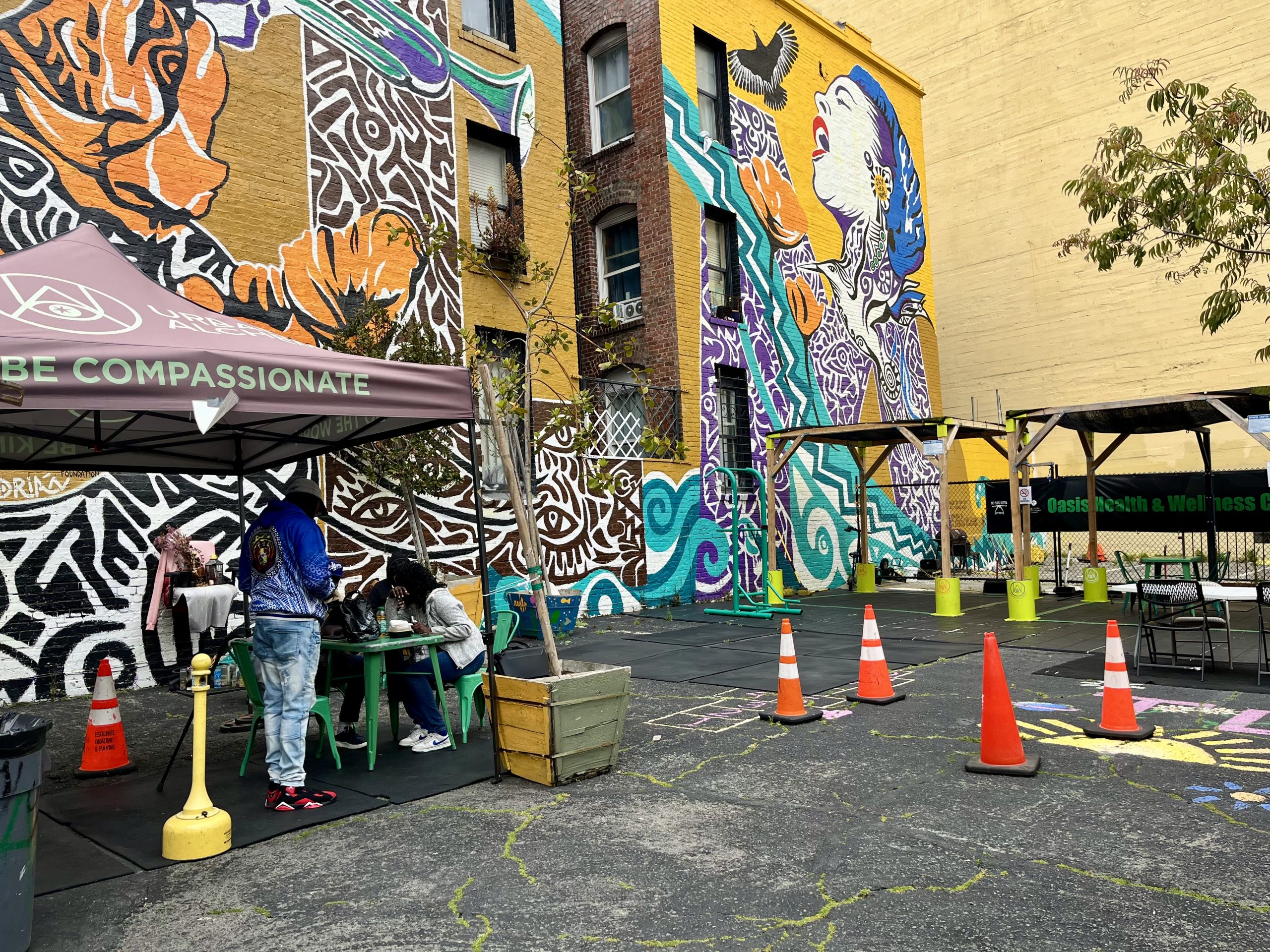 Outdoor urban space with colorful murals, people sitting under a canopy labeled "BE COMPASSIONATE," traffic cones, and planters on a paved lot next to a brick building.
