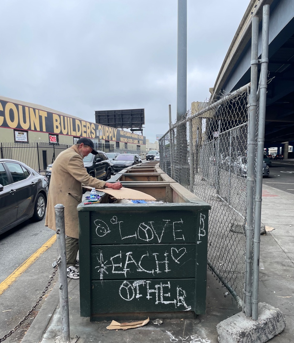 A person in a coat stands by a green dumpster with "LOVE EACH OTHER" written in chalk, next to a chain-link fence and a street under an overcast sky.