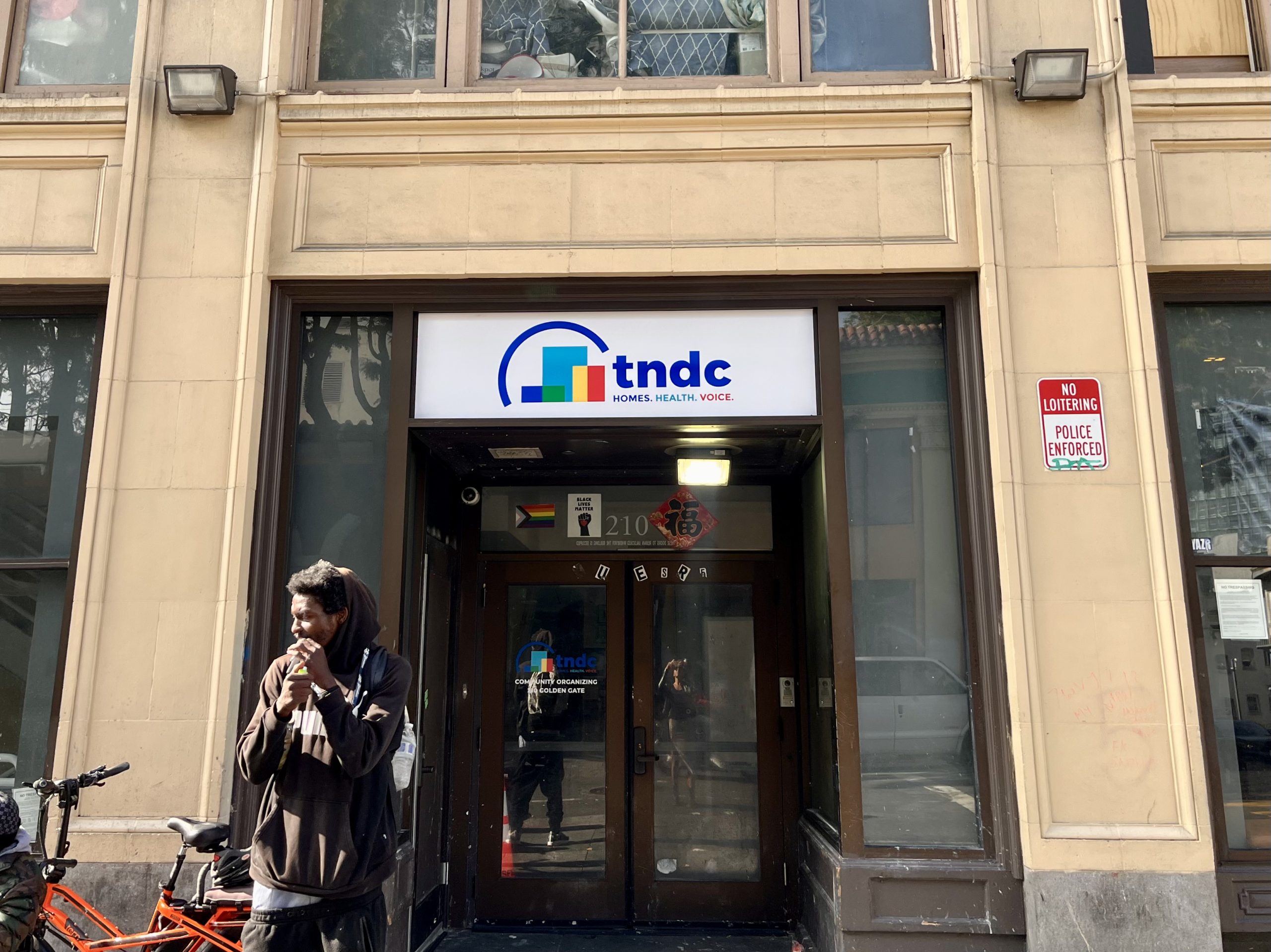 A man stands outside the entrance of a building with a "TNDC: Homes, Health, Voice" sign above the door and a "No Loitering Police Enforced" sign on the wall.