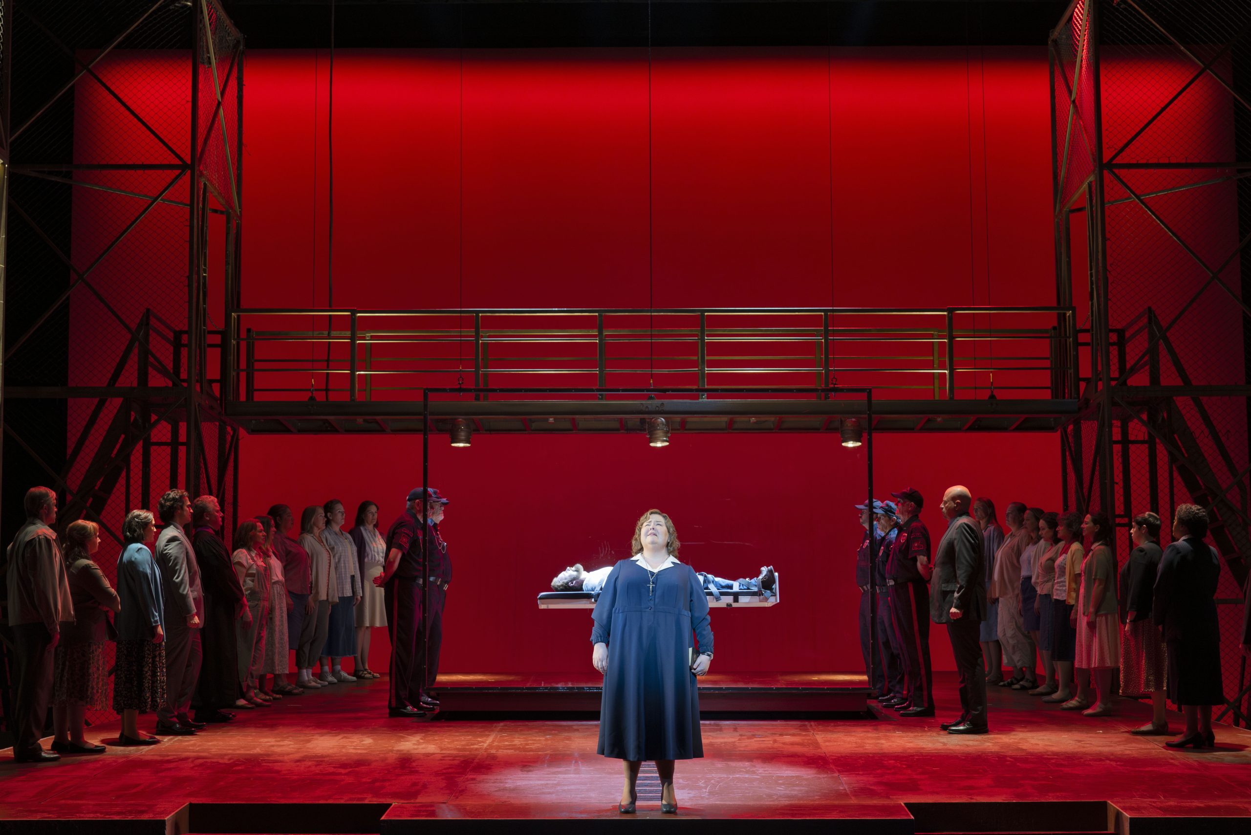 A woman stands center stage in front of a lit platform with a person lying down, surrounded by uniformed guards and two groups of onlookers, set against a vivid red backdrop.