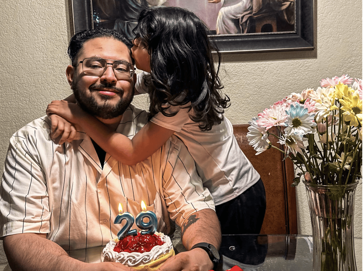 A man sits at a table with a small birthday cake showing the number 29, while a child hugs and whispers to him. A vase of flowers is on the table.