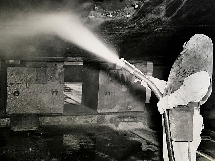 A worker wearing protective gear uses a hose to sandblast a large metal surface in an industrial setting.