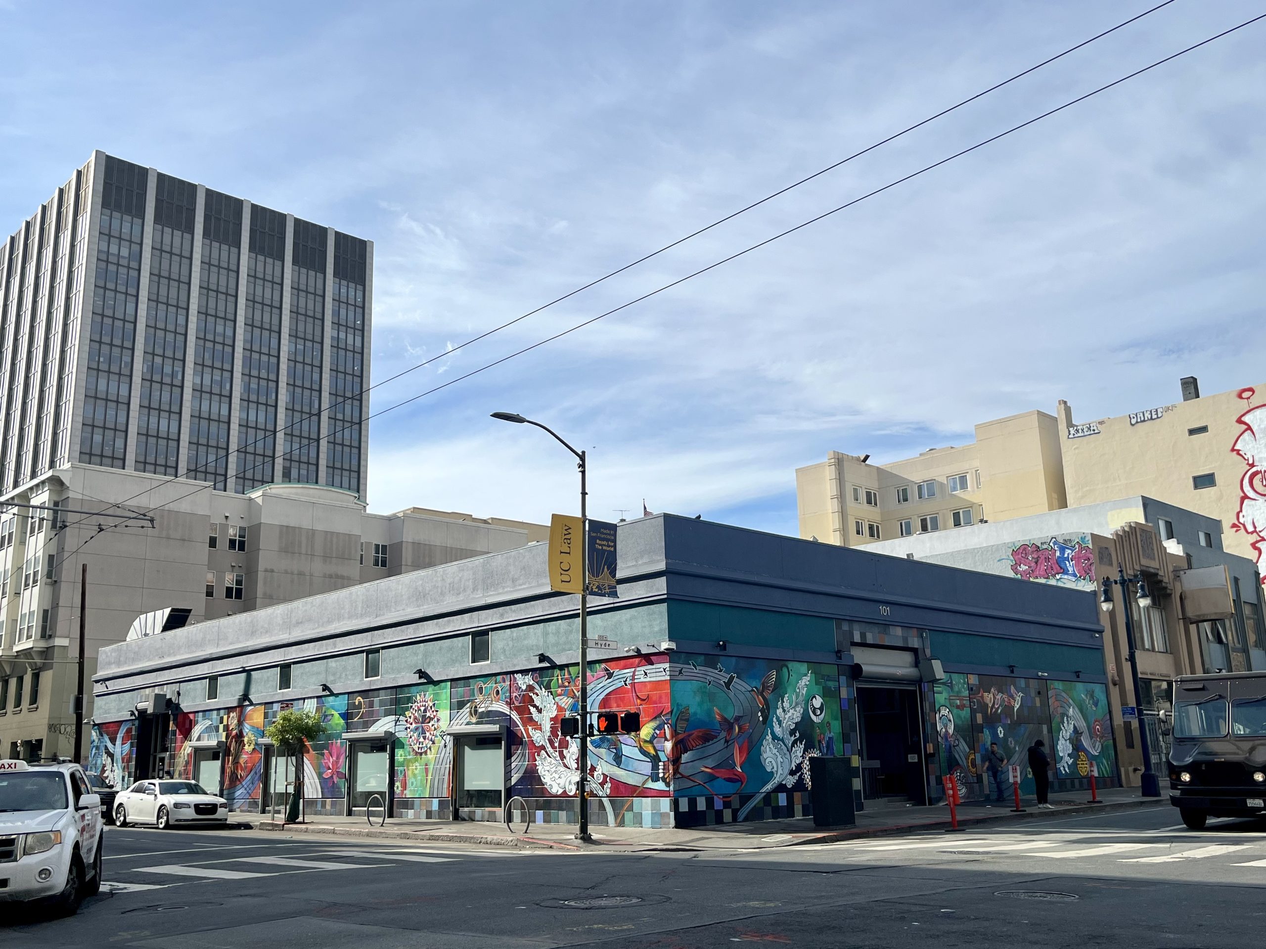 A single-story building on a city corner features colorful murals on its walls, with cars and tall buildings visible in the background under a partly cloudy sky.
