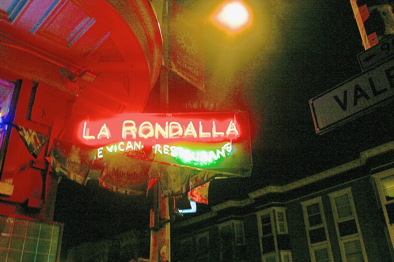 Neon sign for "La Rondalla Mexican Restaurant" glows at night on a street corner, with a streetlight and "Valencia" street sign visible.