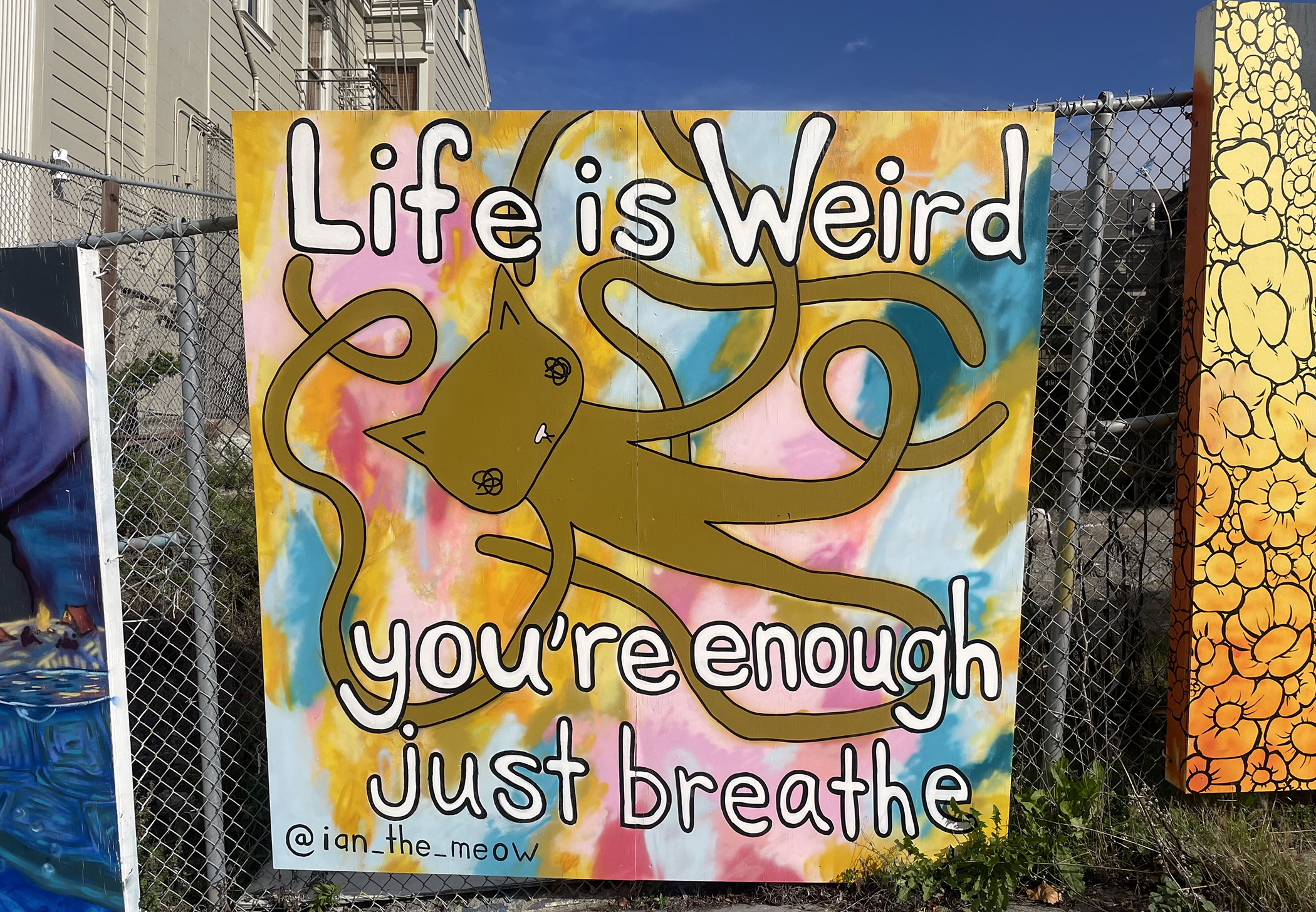 A mural features a cat with wavy tentacles and the text “Life is Weird, you’re enough, just breathe” against a colorful background.