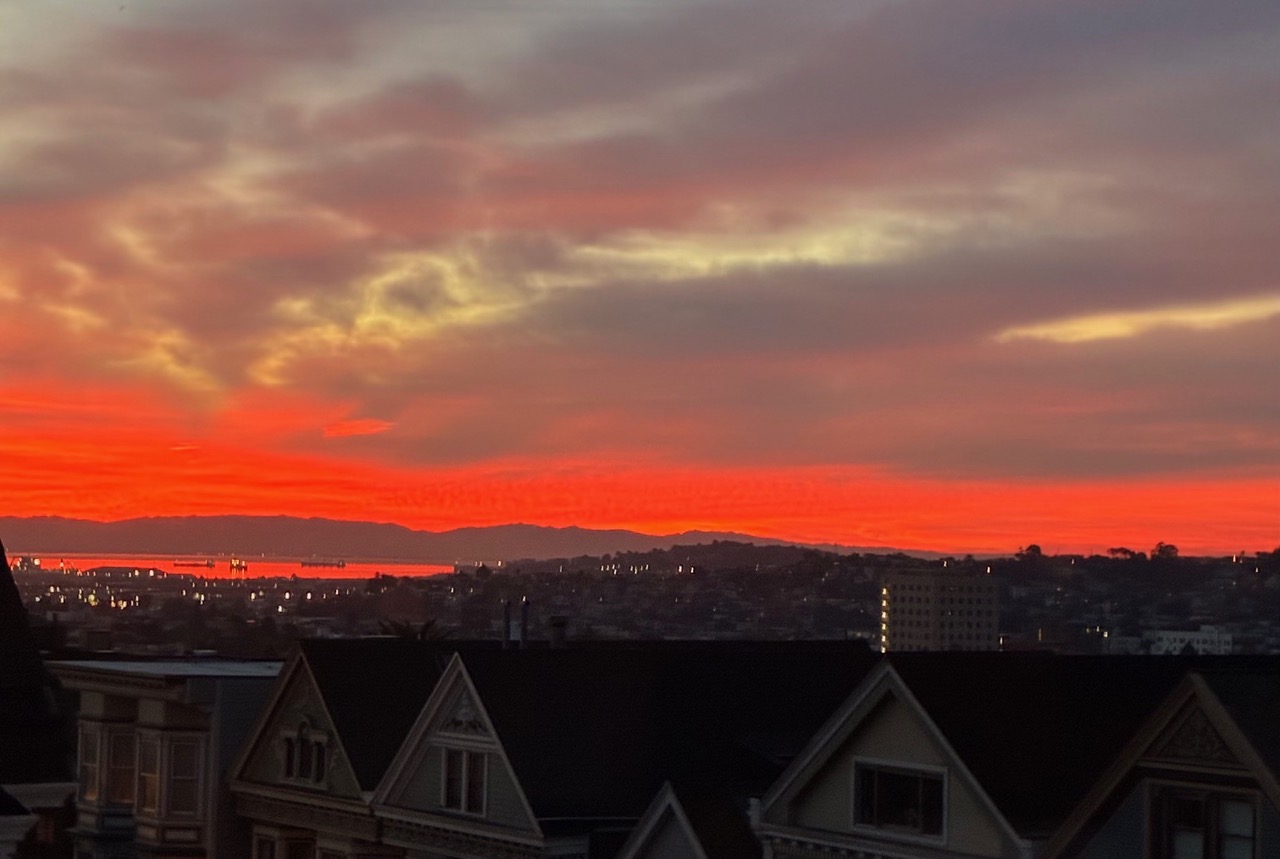 Fiery orange and red sunset sky over a cityscape with silhouetted rooftops and distant hills.