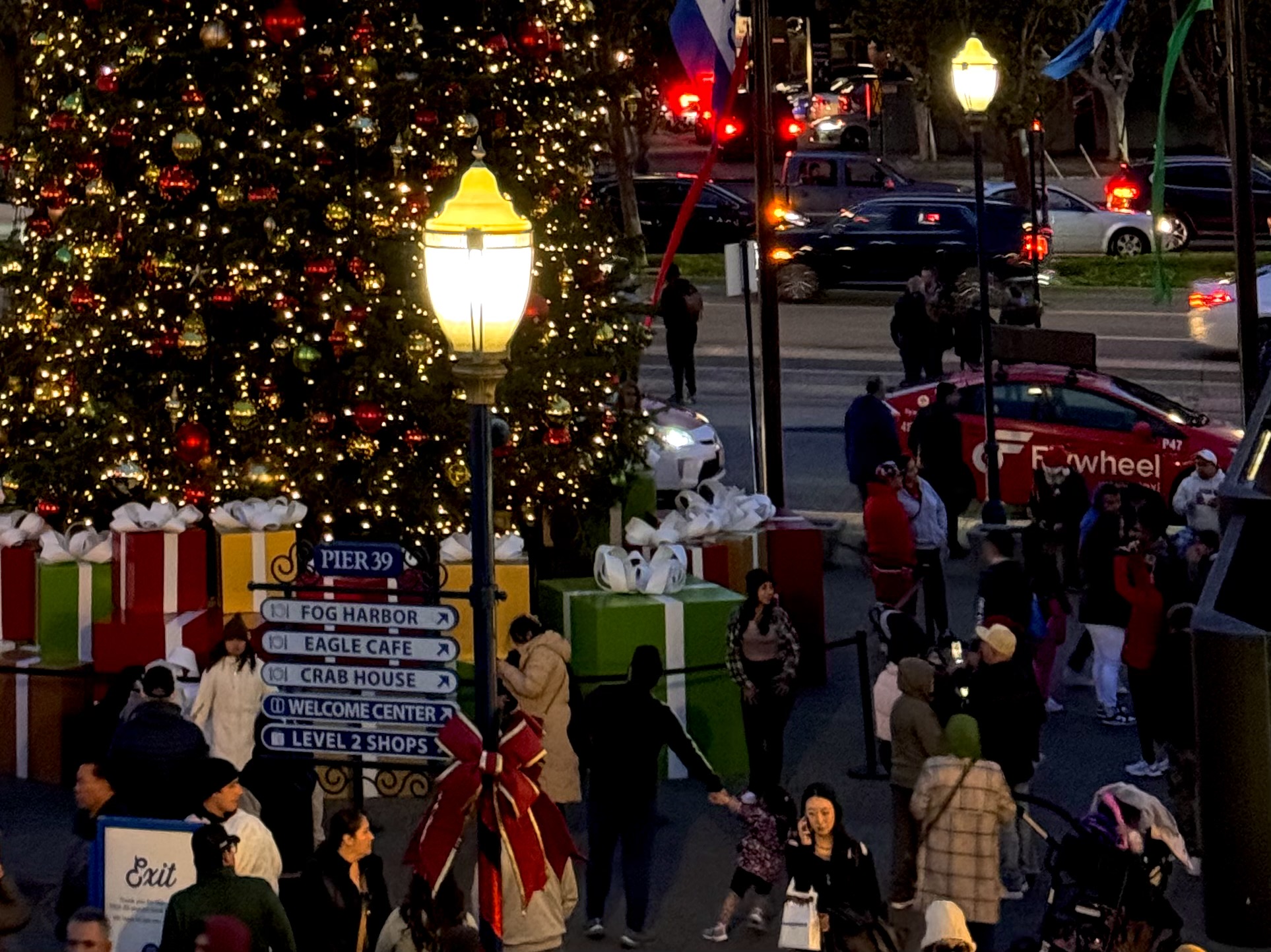 A decorated Christmas tree with sparkling lights and gift boxes stands near a directional sign at Pier 39 as people stroll around and cars drive by in the festive background.