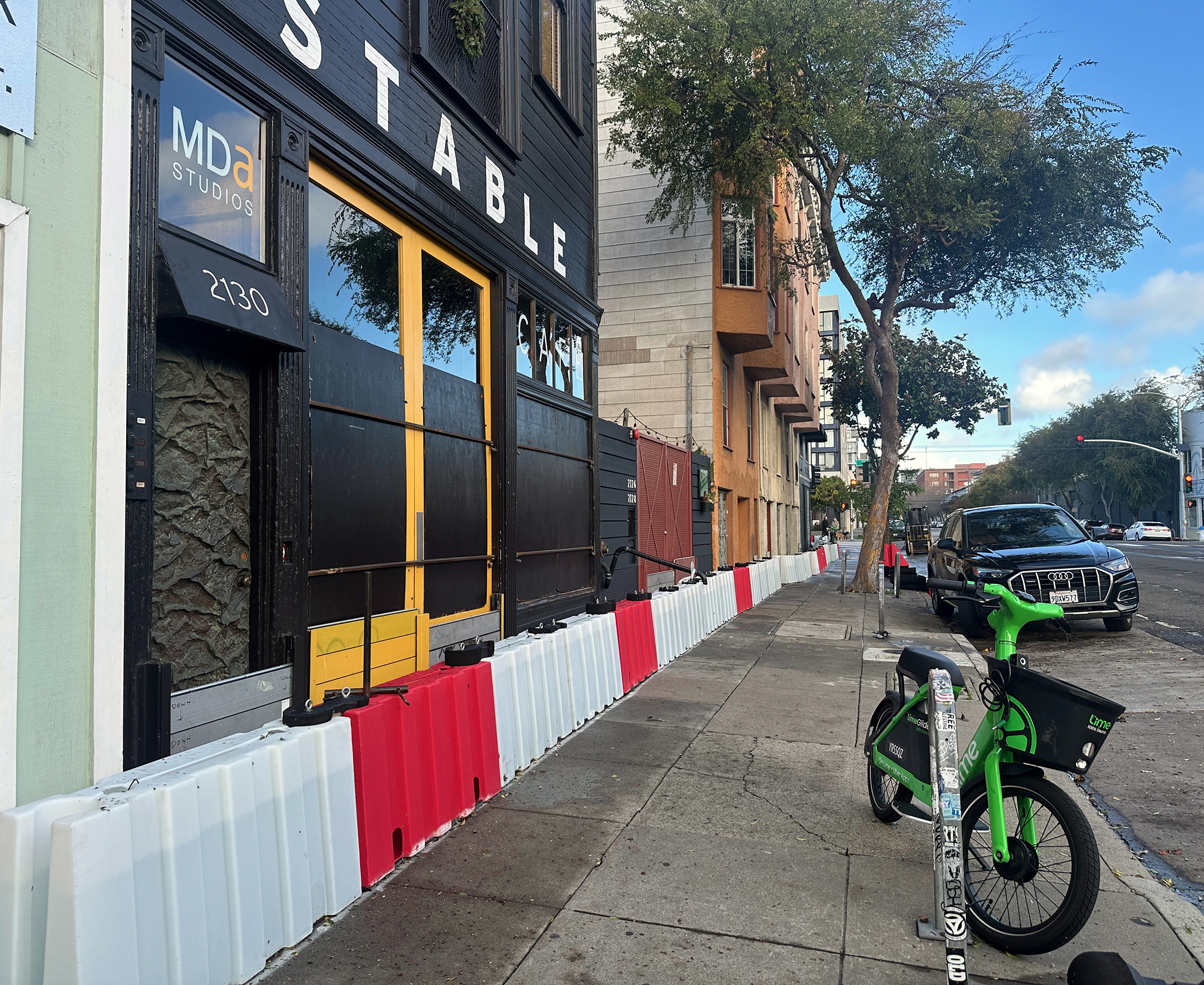 A sidewalk lined with red and white barricades next to a building with yellow doors; a parked green rental bike and a black car are visible on the right.