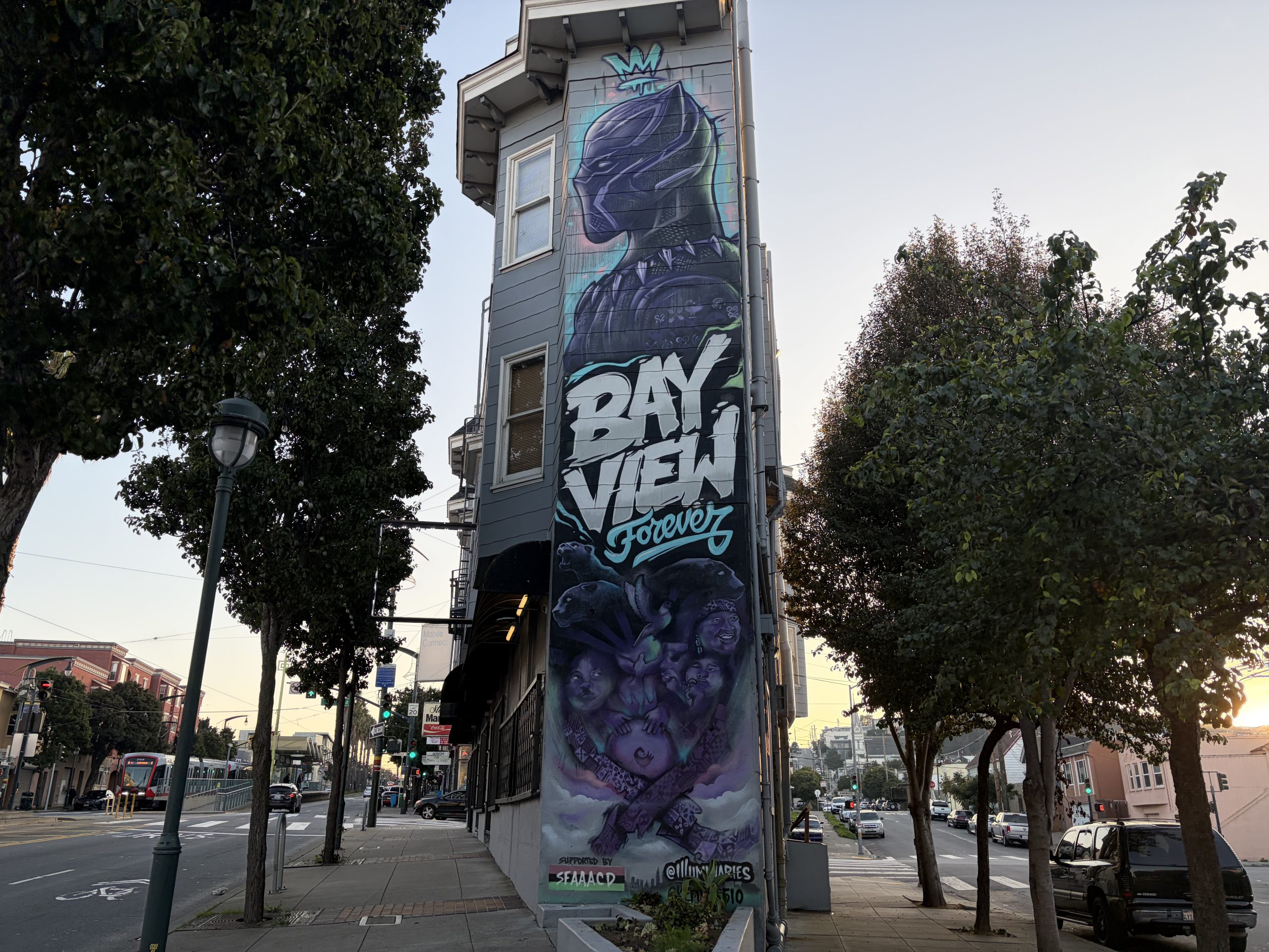 A tall, narrow building features a mural of Marvel’s Black Panther, the text “Bayview Forever,” and faces of children painted below, located on a city street corner at sunset.