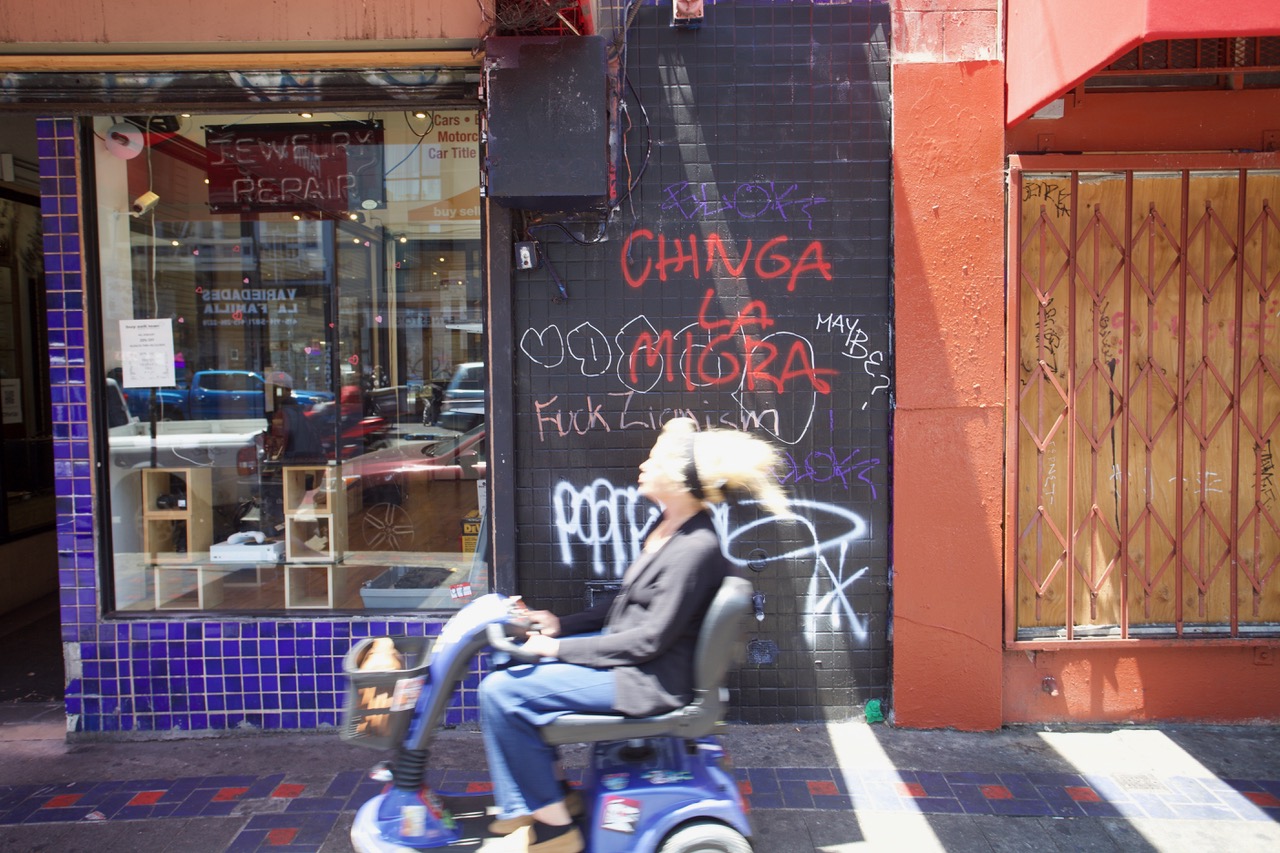A person rides a mobility scooter past a wall with graffiti and a boarded-up storefront in an urban setting.