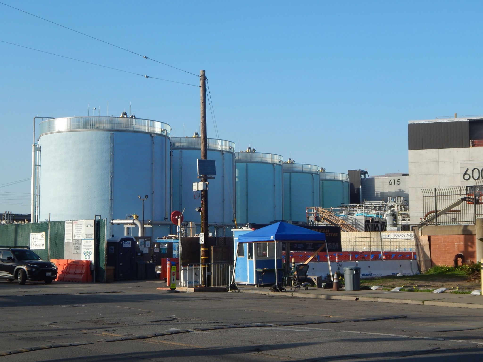 Large cylindrical industrial storage tanks are situated behind a gate and a small blue security booth, with various industrial equipment and buildings visible nearby.