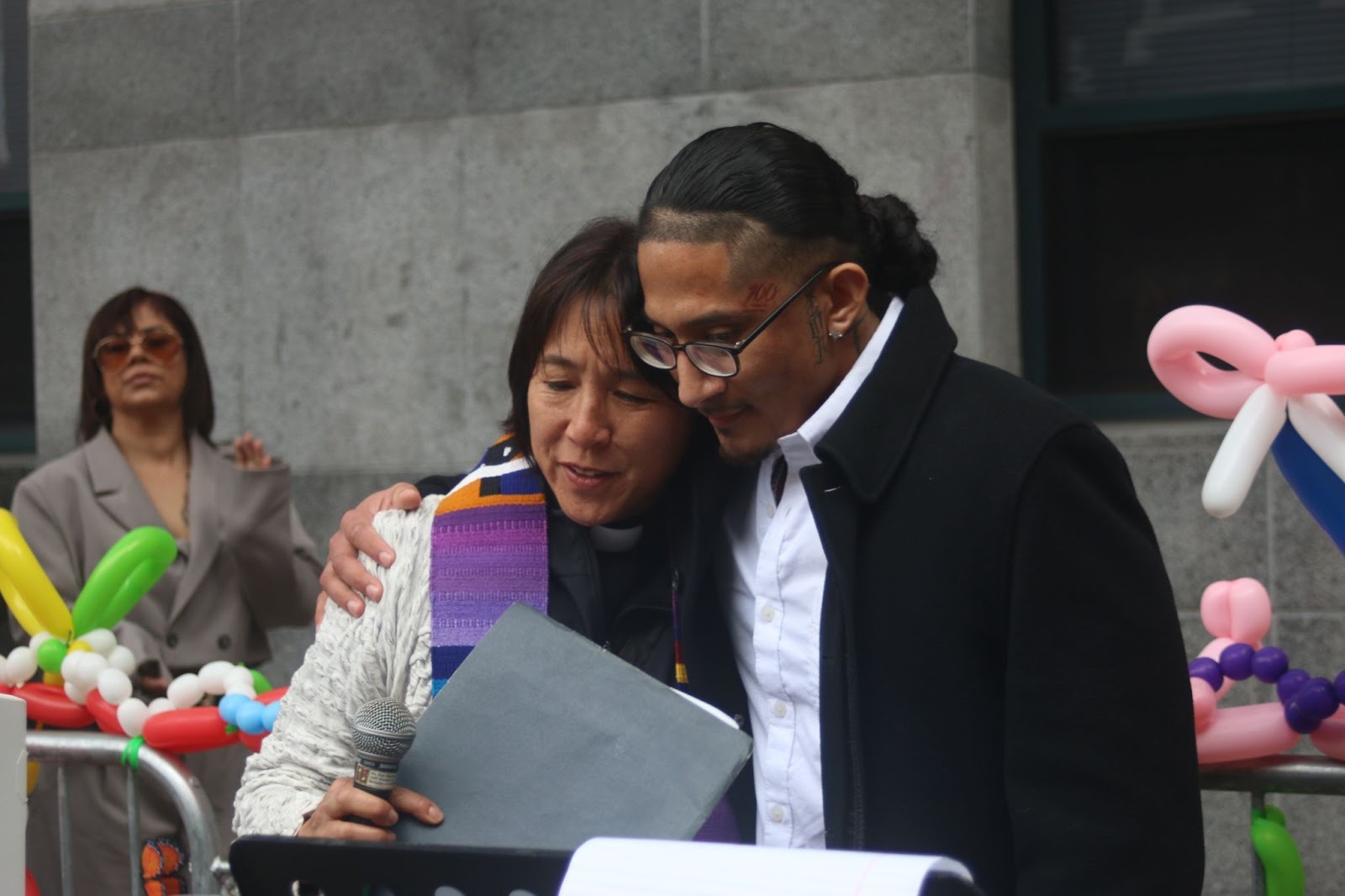 Two people stand close together, one with an arm around the other, holding papers. Balloons decorate the background, and another person stands behind them.