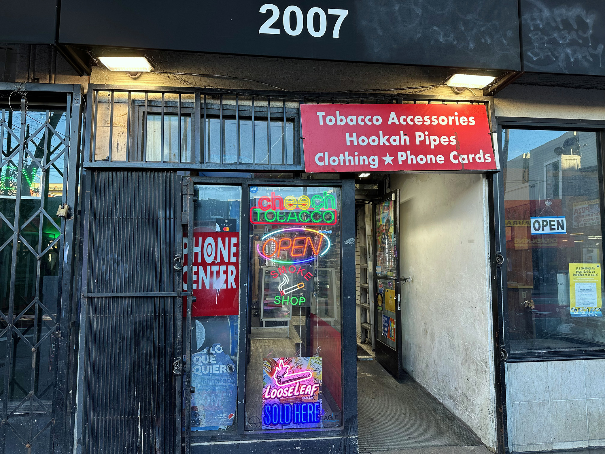 Storefront with barred window and door, neon signs for tobacco and phone cards, and a red sign above reading "Tobacco Accessories, Hookah Pipes, Clothing, Phone Cards.