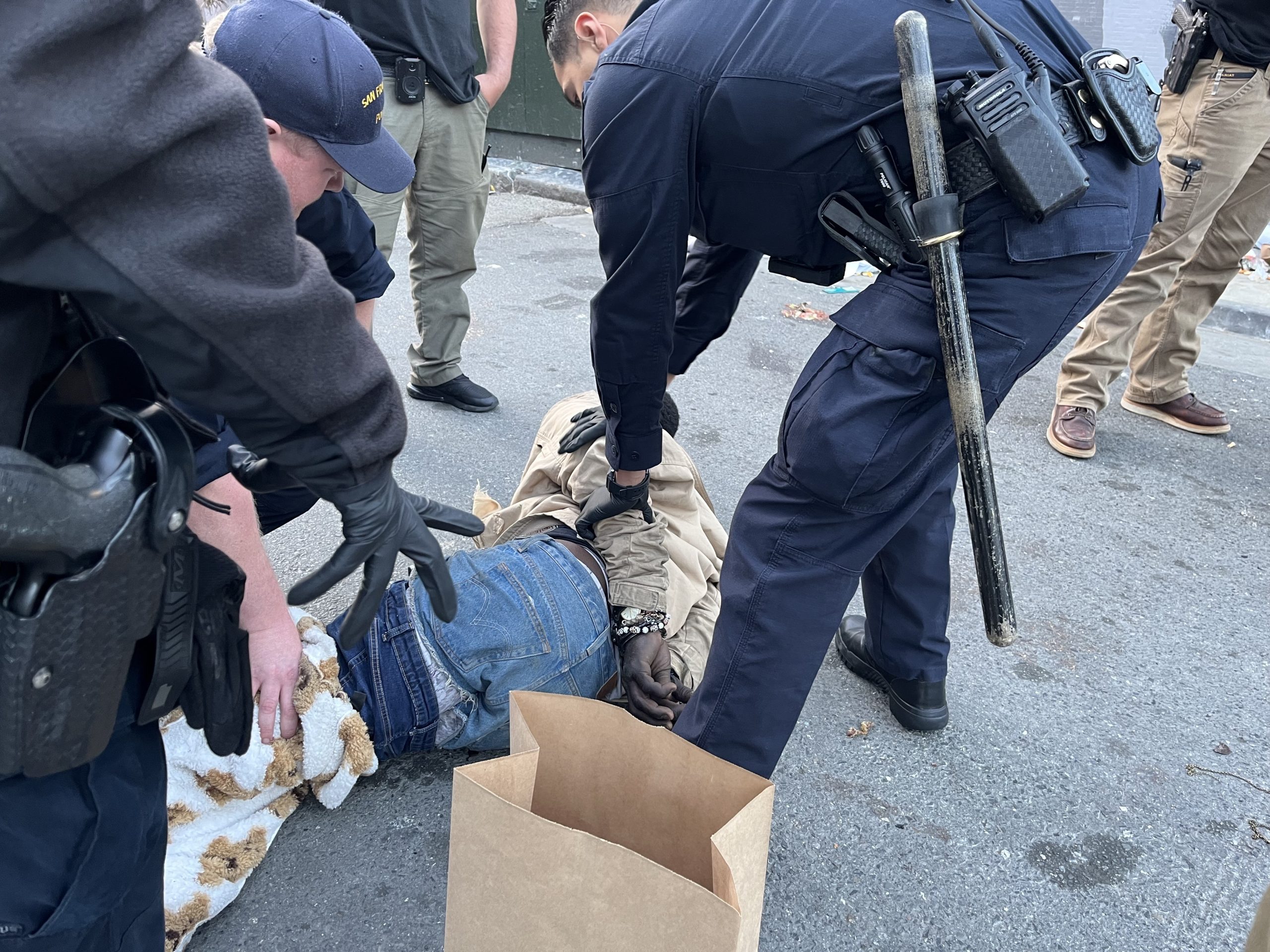 Police officers detain a person lying on the ground as they restrain and handcuff him on a street near a cardboard box and a blanket.