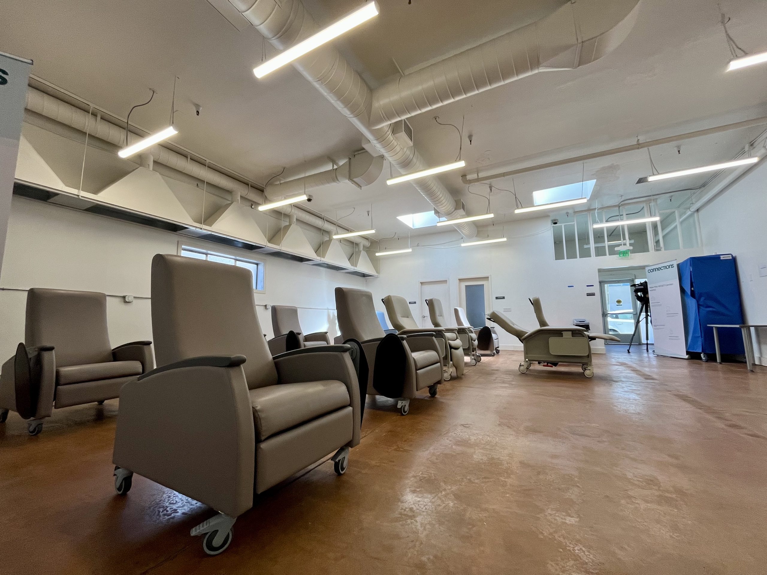A room with several gray reclining chairs on wheels arranged in rows, fluorescent ceiling lights, and medical equipment visible in the background.