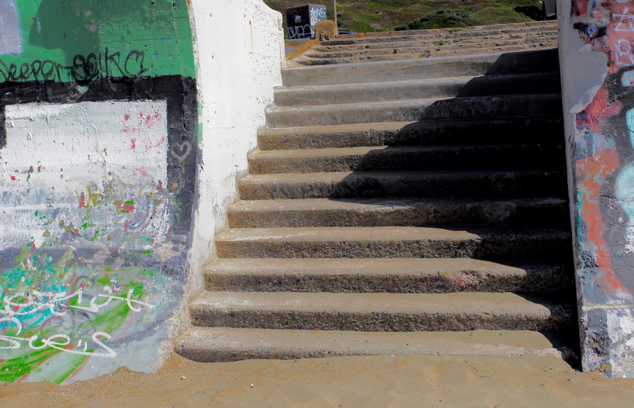 Concrete outdoor steps covered in sand lead upward beside a graffiti-covered wall, with sunlight casting shadows across the scene.