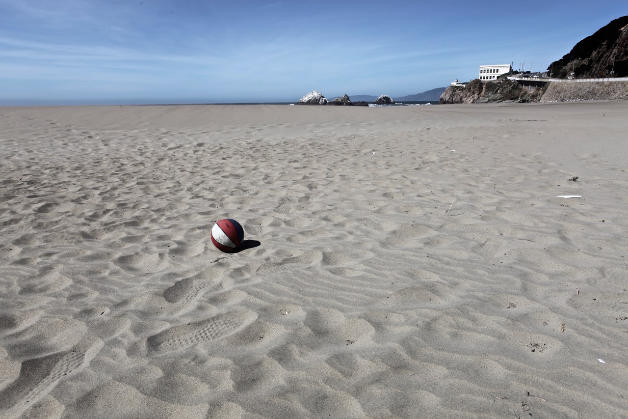 A red and white basketball sits alone on a sandy beach under a clear sky, with distant rocks and a building visible near the shoreline.