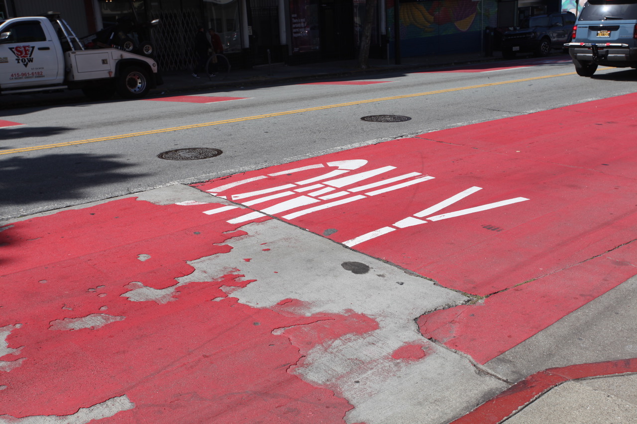 Red bus lane on a city street with faded paint and "ONLY" visible in white letters; cars parked nearby and a tow truck in the background.
