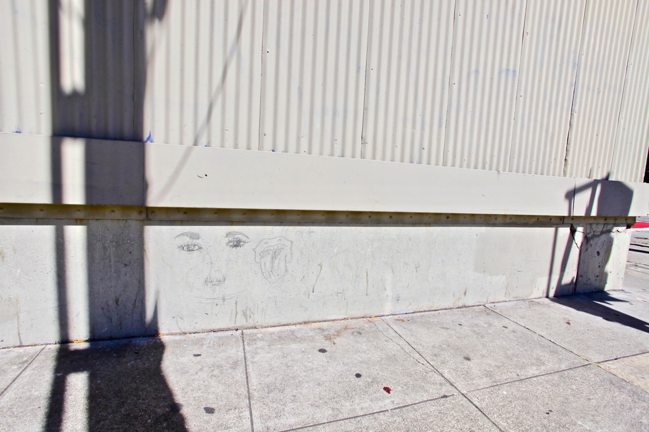 Faint pencil sketch of a face and a flower on a concrete wall beneath a metal railing, with shadows cast across the sidewalk and wall.