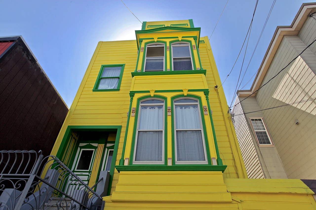A two-story yellow house with green trim, featuring arched windows with white curtains and a front entry door, viewed from a low angle.