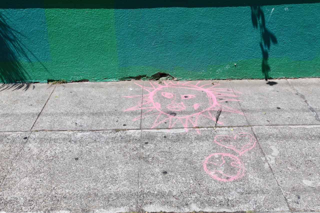 Pink chalk drawing of a sun with a face, a heart, and a peace symbol on a sidewalk next to a green painted wall.