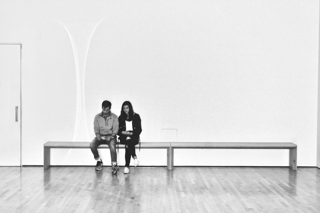 Two people sit next to each other on a long wooden bench against a plain white wall in a mostly empty room with wooden flooring.
