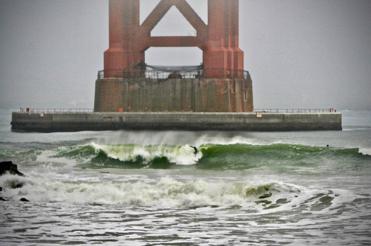 A surfer rides a wave near the base of a large red bridge structure on a foggy day, with choppy water and concrete barriers visible.