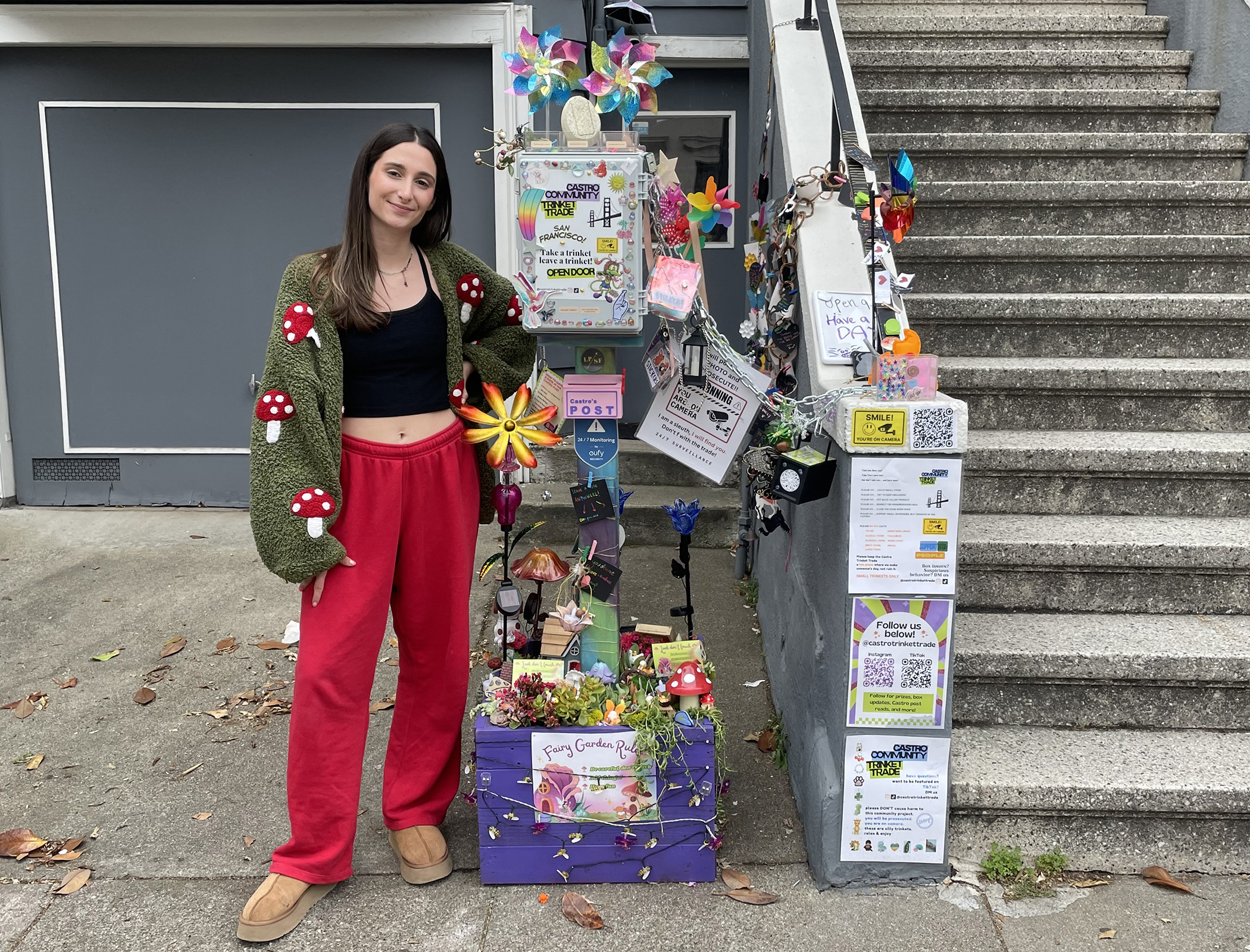 A woman in red pants and a green sweater with mushroom designs stands next to a colorful decorated display box by outdoor steps. The display features flowers, flyers, and crafts.
