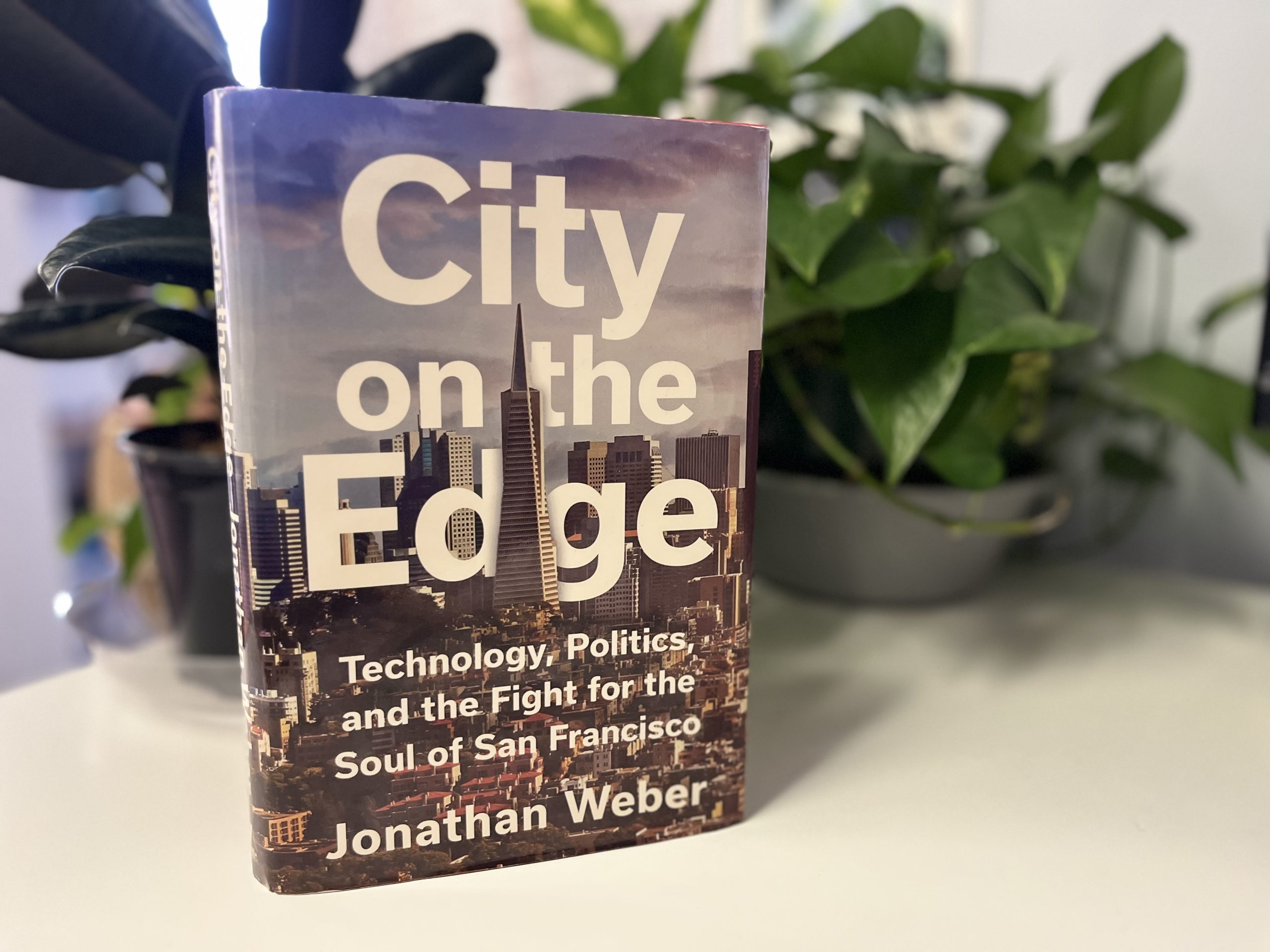 The book "City on the Edge" by Jonathan Weber stands upright on a white surface, with leafy green plants in the background.