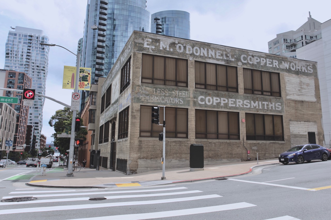 A weathered two-story building with faded signs for "E.M. O'Donnell Copper Works" and "Coppersmiths" stands at a city intersection surrounded by modern high-rise buildings.