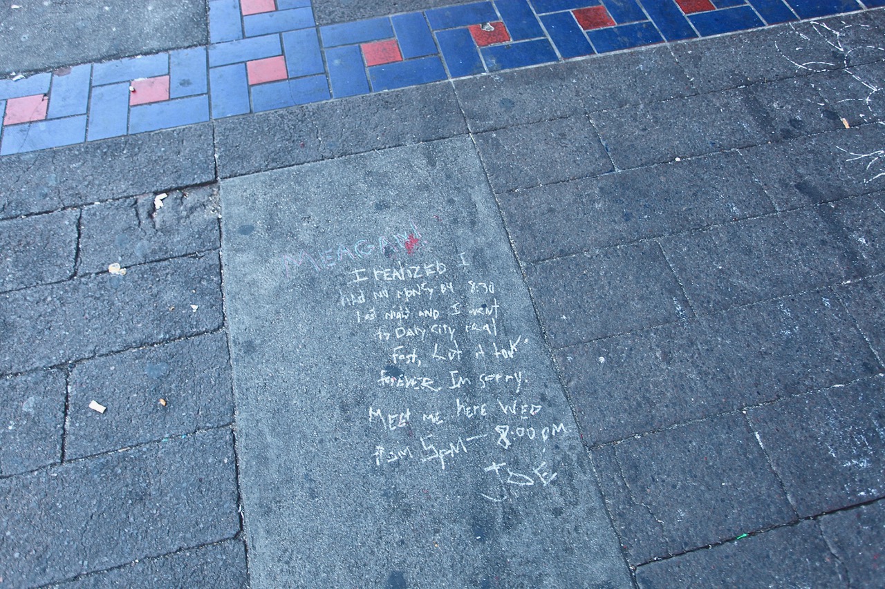 Chalk-written message on a sidewalk, partially legible, with bricks in red and blue patterns nearby. The note includes an apology, a time, and is signed "Joe.