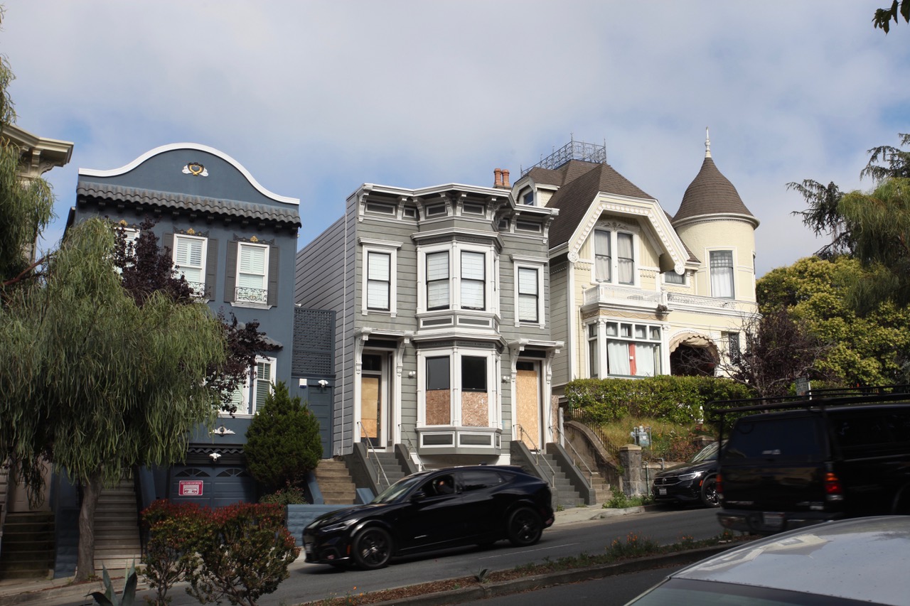 Three Victorian-style houses line a street; the center house has boarded-up windows, and cars are parked along the curb under a partly cloudy sky.