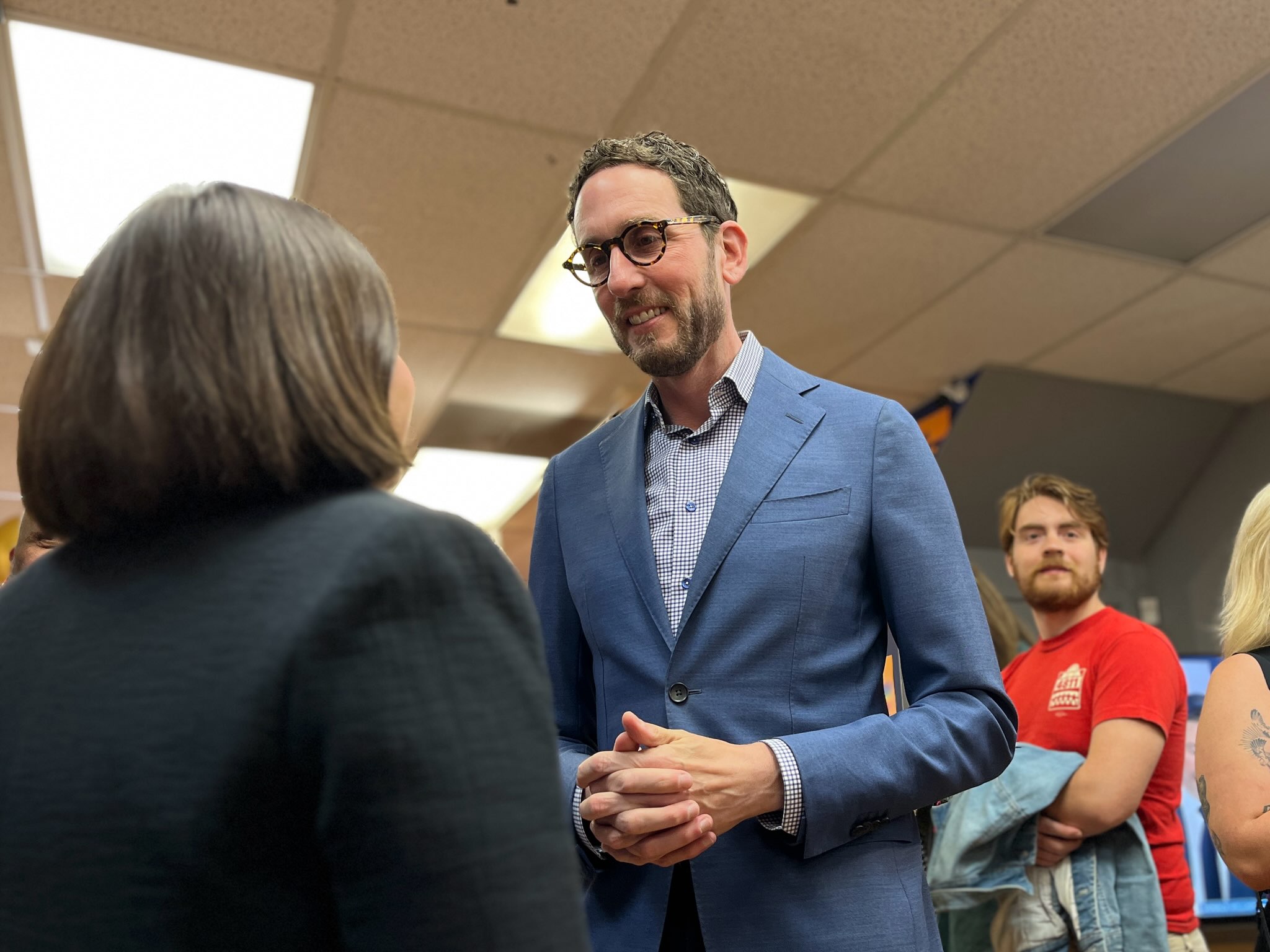 A man in a blue suit speaks to a woman in a black jacket at an indoor event while other people stand in the background.