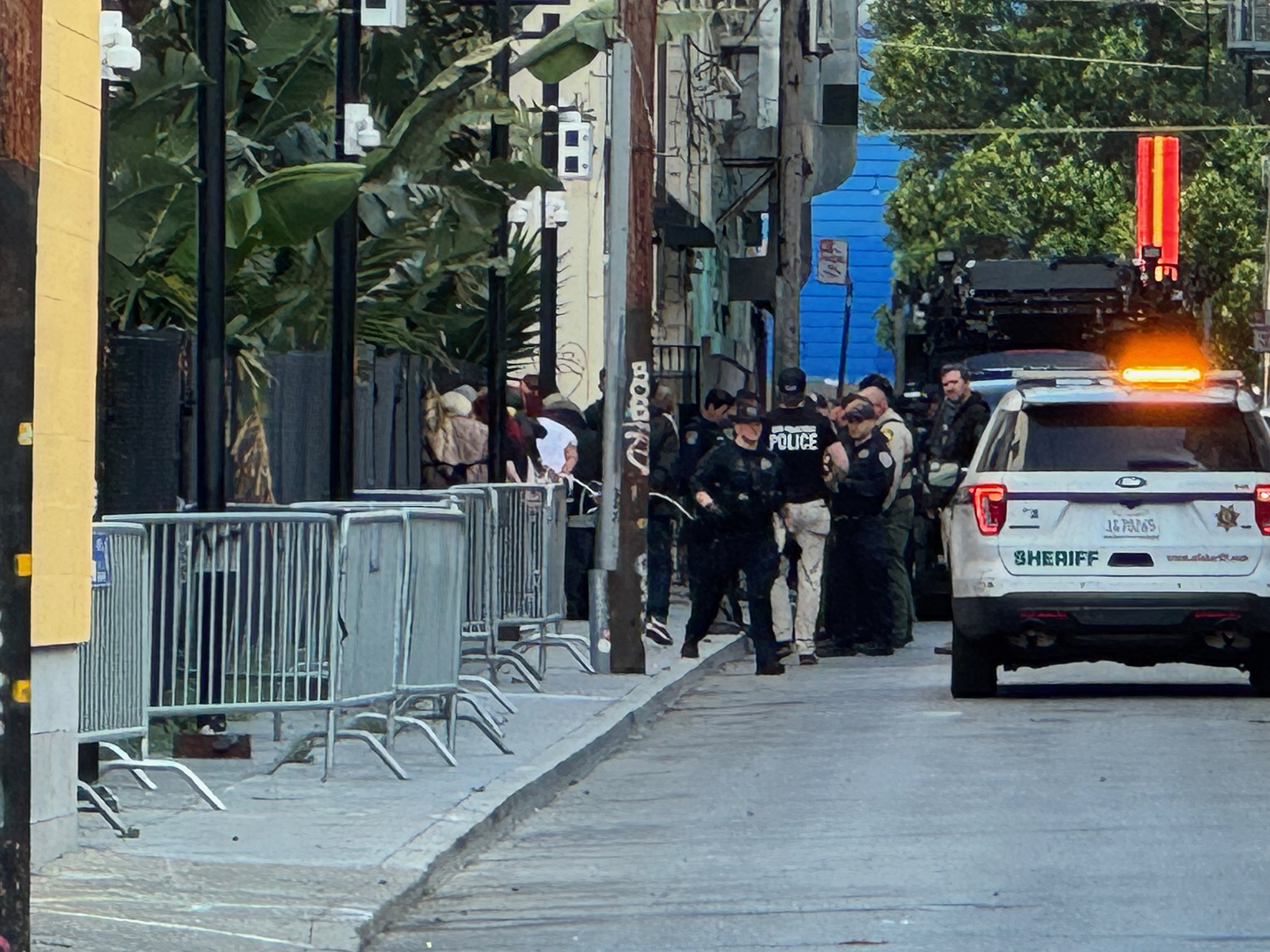 Police officers gather near a sheriff’s vehicle on a city street, with metal barricades and people lined up along the sidewalk in the background.