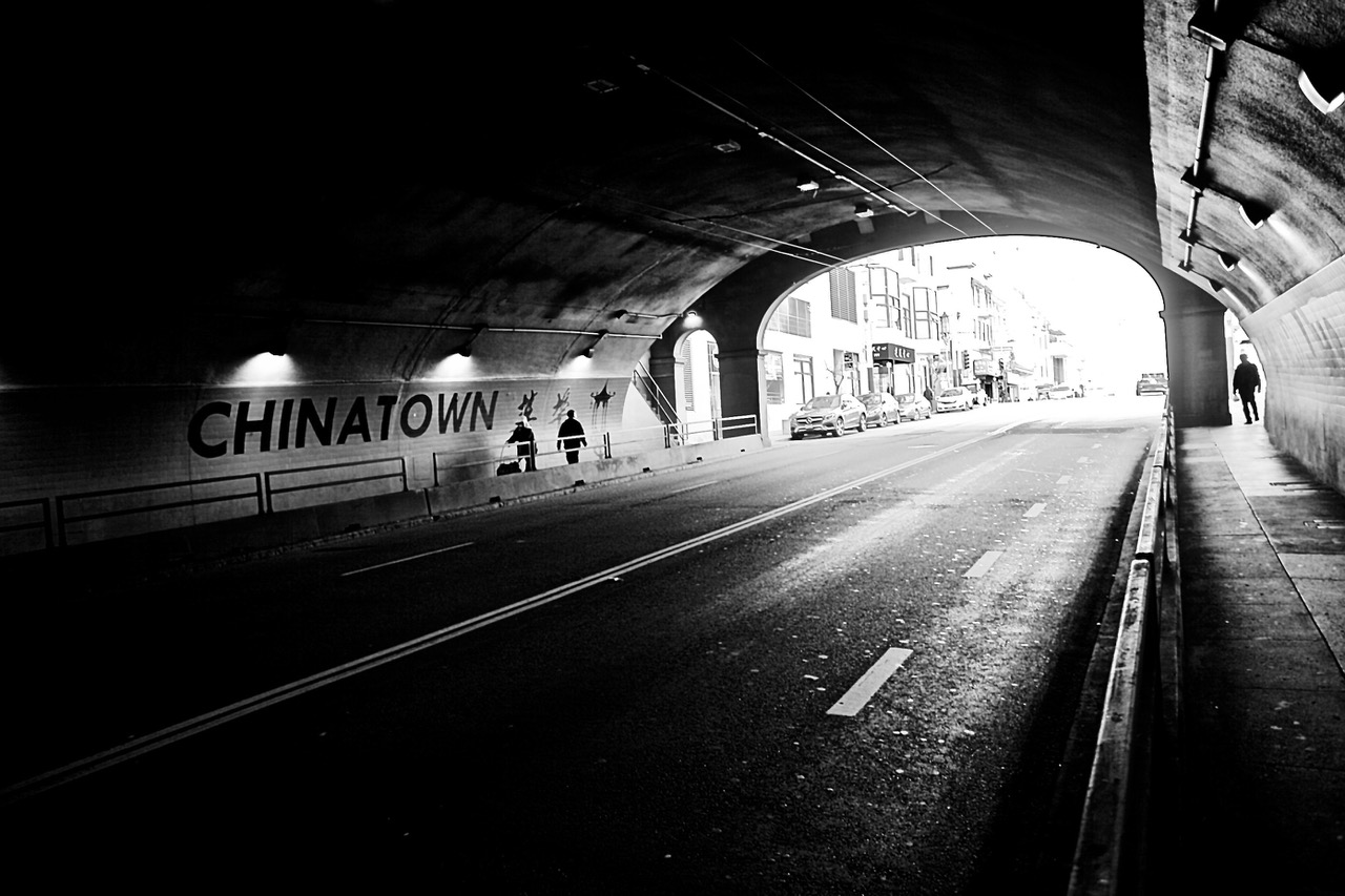 Black and white photo of a tunnel with "CHINATOWN" painted on the wall, leading to a city street with parked cars and a lone person walking on the sidewalk.