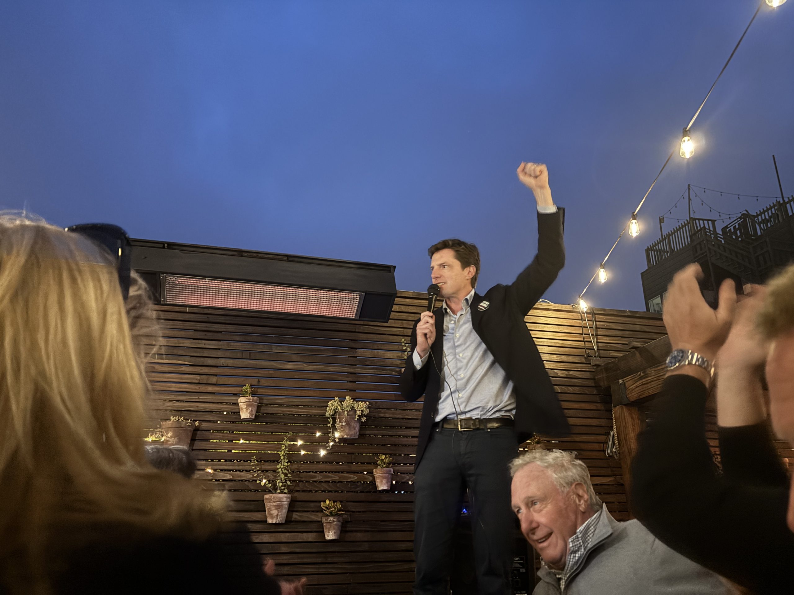 A man stands on a bench holding a microphone with one fist raised, addressing a small crowd at an outdoor venue with string lights and a wooden wall decorated with potted plants.