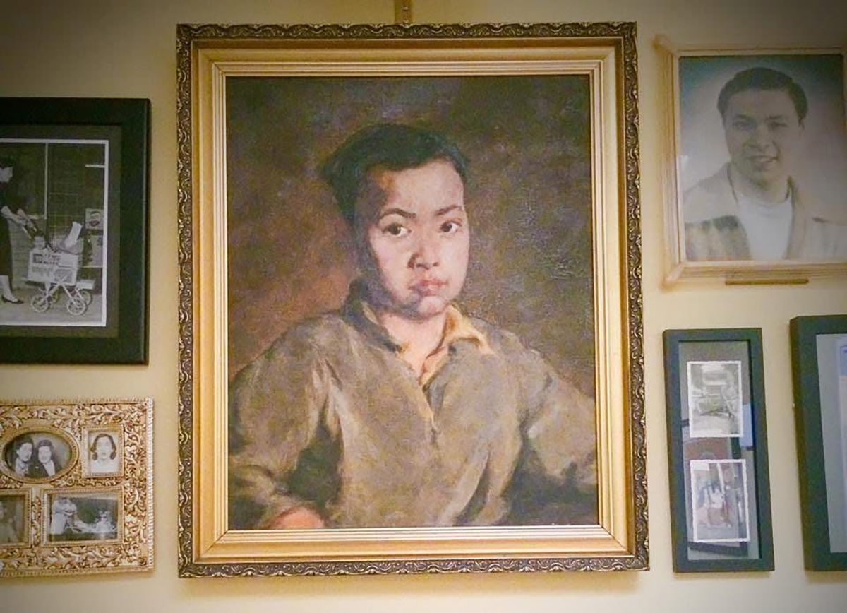 A framed portrait painting of a young man is centered on a wall, surrounded by various framed photographs and picture collages.