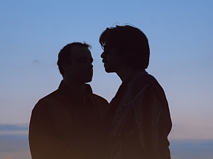 Two people stand facing each other in silhouette against a blue and orange sky at dusk.