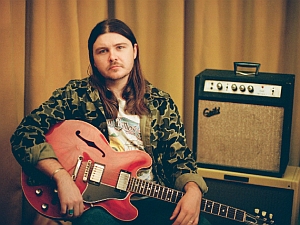 Man with long hair sits holding a red electric guitar in front of a beige curtain, next to a guitar amplifier.