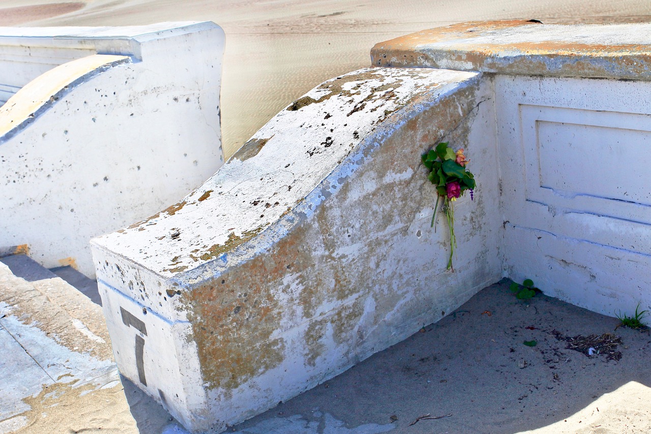 Weathered white concrete structure by a sandy beach with wilted pink and yellow flowers placed in a crevice.
