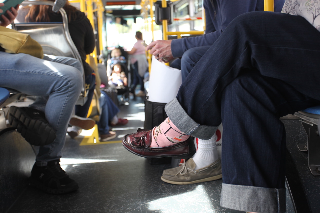 People sitting on a bus; foreground shows crossed legs wearing pink socks and loafers. The bus interior and other riders are visible.