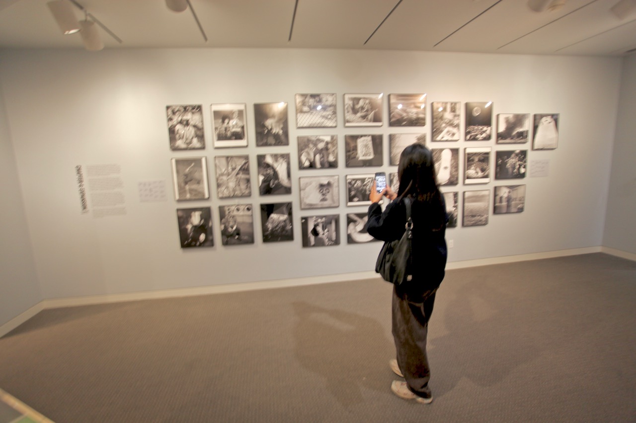 A person stands in an art gallery photographing a wall display of black-and-white photographs arranged in a grid.