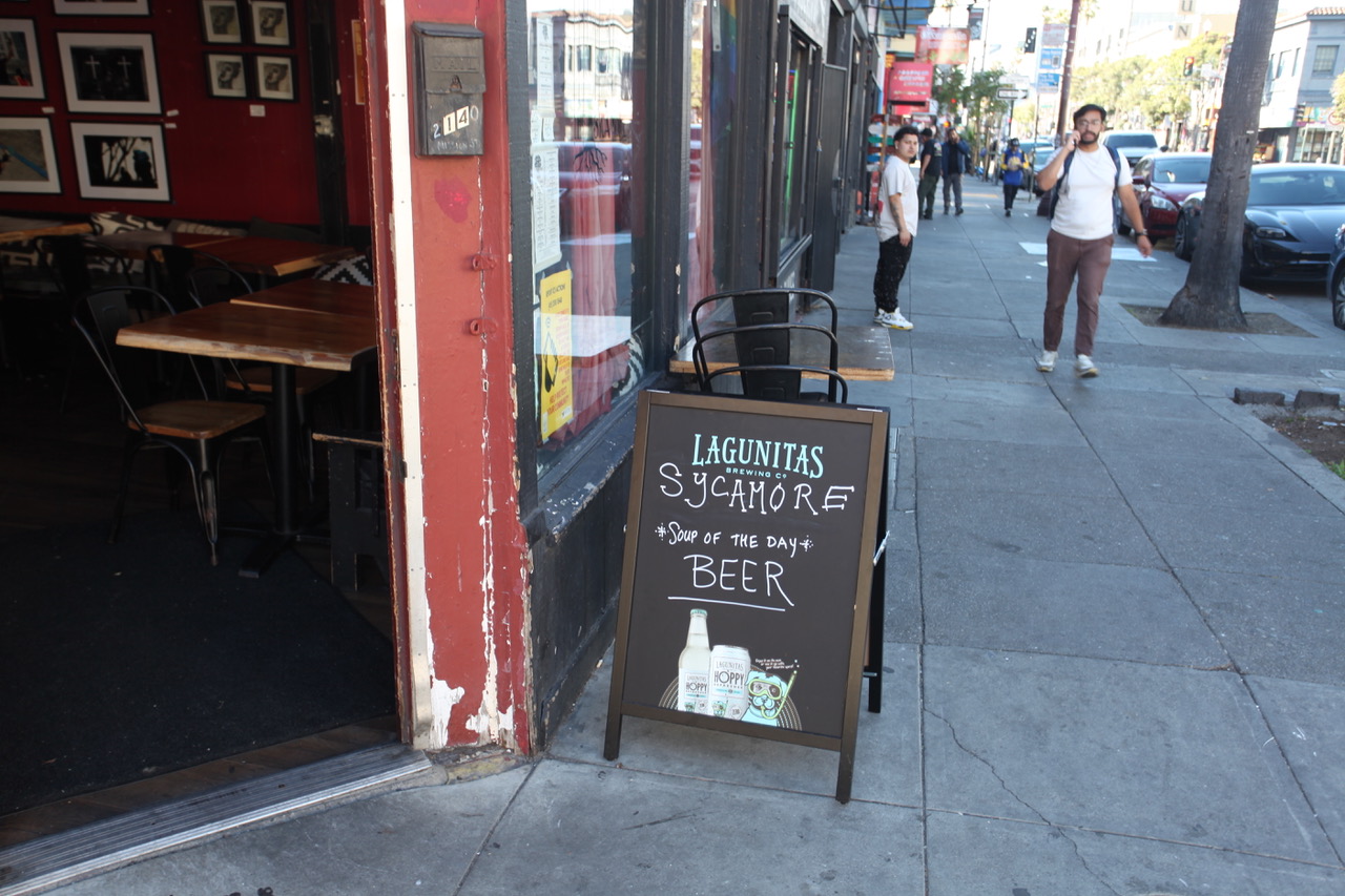 A sidewalk chalkboard outside a restaurant advertises "Lagunitas Sycamore Soup of the Day: Beer." Several people walk along the street.