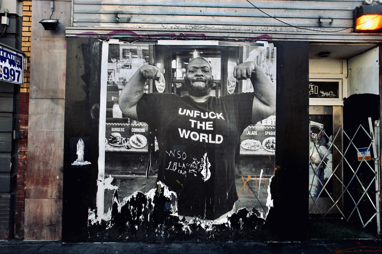 A large black-and-white mural shows a man flexing his arms and wearing a shirt that says “UNFUCK THE WORLD.” The bottom of the mural is torn and has graffiti.